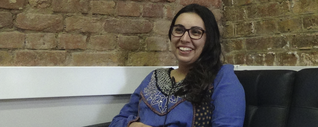 A imagem mostra uma mulher branca, de cabelos escuros, usando óculos, sorrindo e vestindo uma blusa azul. Está sentada em um sofá preto em frente a uma parede de tijolos