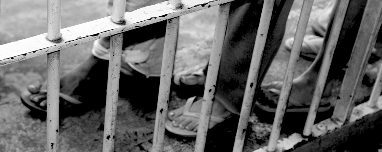 A imagem em preto e branco mostra, atrás de grades, os pés de mulheres calçando chinelos e vestido calças