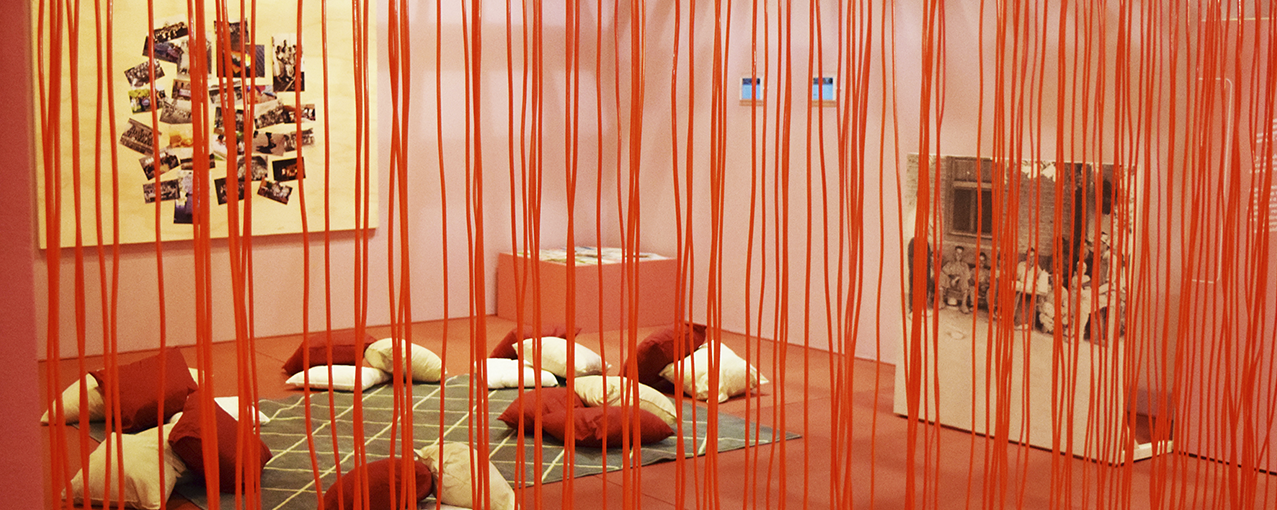 Com pares rosas, a sala conta com uma cortina com fios vermelhos, um tapete e diversas almofadas no chão e fotos em um quadro amarelo claro na parede