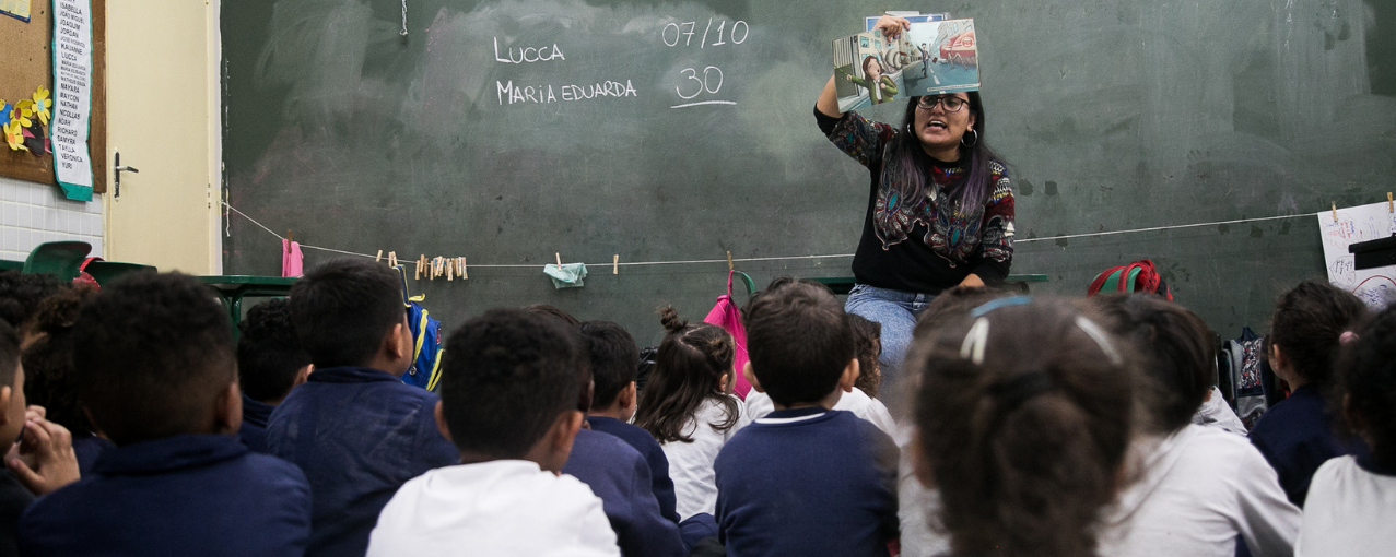 Foto de uma sala de aula com crianças de costas e uma professora.